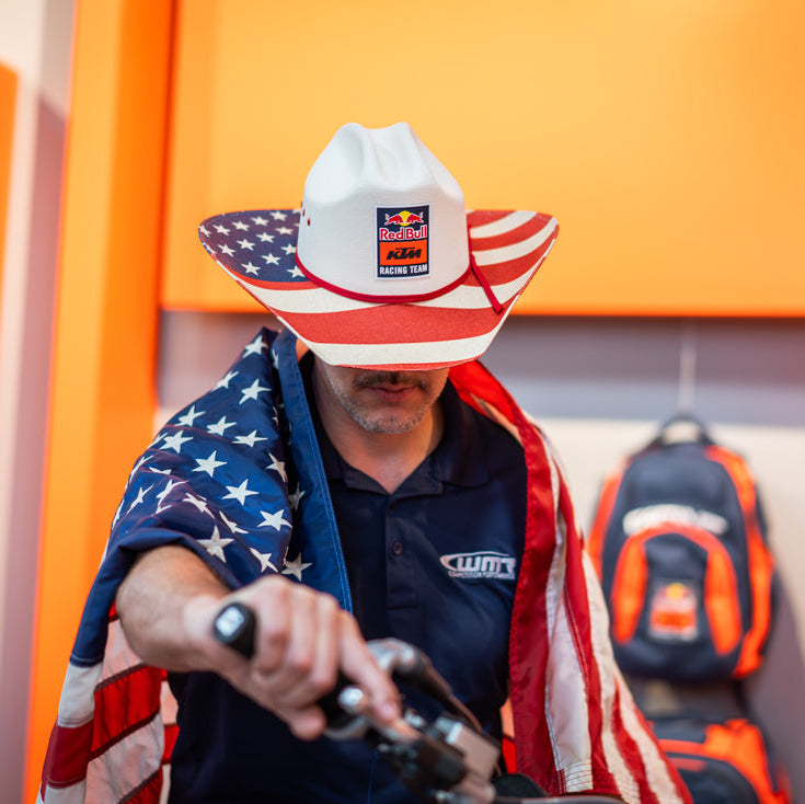 Person wearing a cowboy hat with an American flag design and a branded shirt, standing in front of orange cabinets. Wearing KTM Red Bull Racing Cowboy hat just like AP.