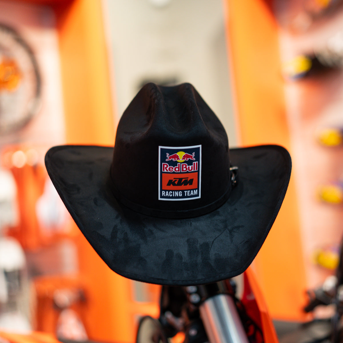 Black cowboy hat with Red Bull KTM Racing Team logo on a blurred background of orange and white elements.